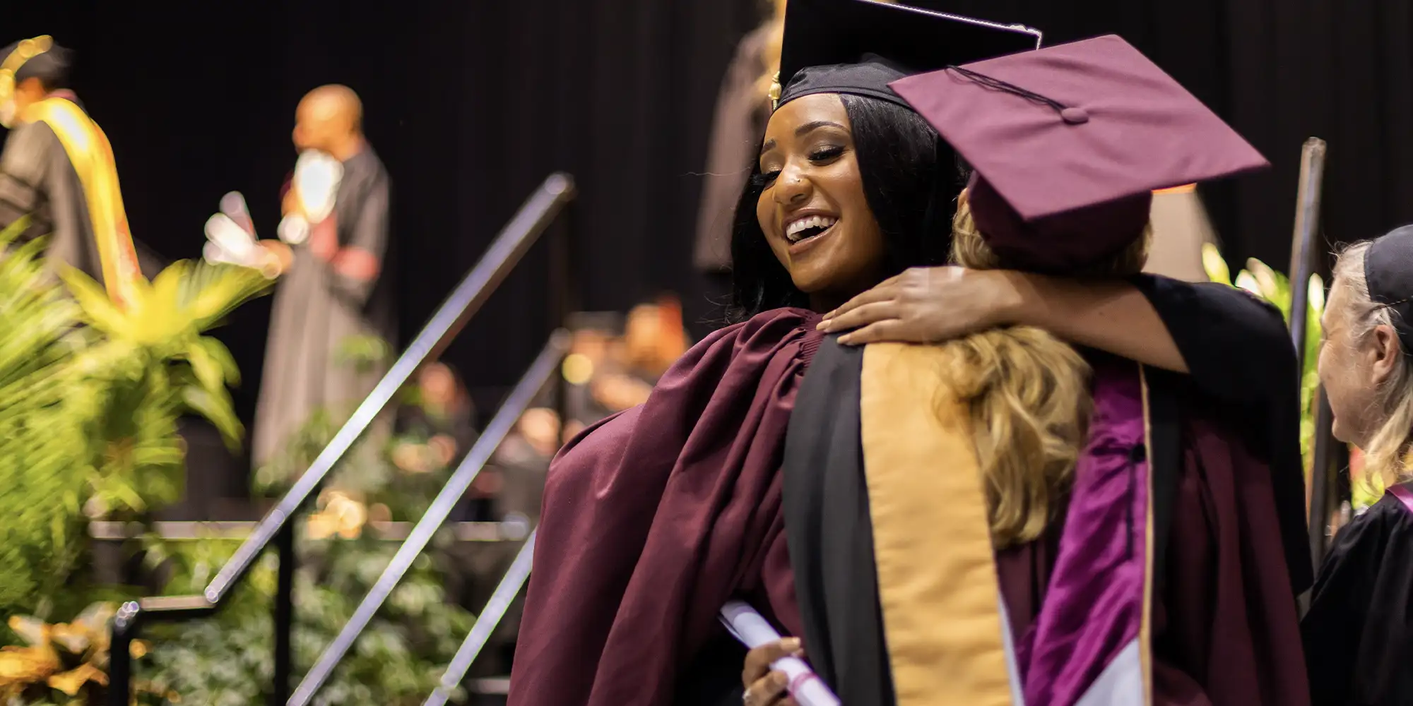 A joyful graduate in a maroon cap and gown embraces another graduate during a commencement ceremony. She is smiling while holding a diploma, surrounded by faculty and other graduates in academic regalia on stage.